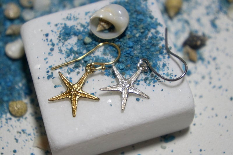 A pair of gold starfish earrings with silver star-shaped pendants, placed on a white surface with blue sand and seashells scattered around.