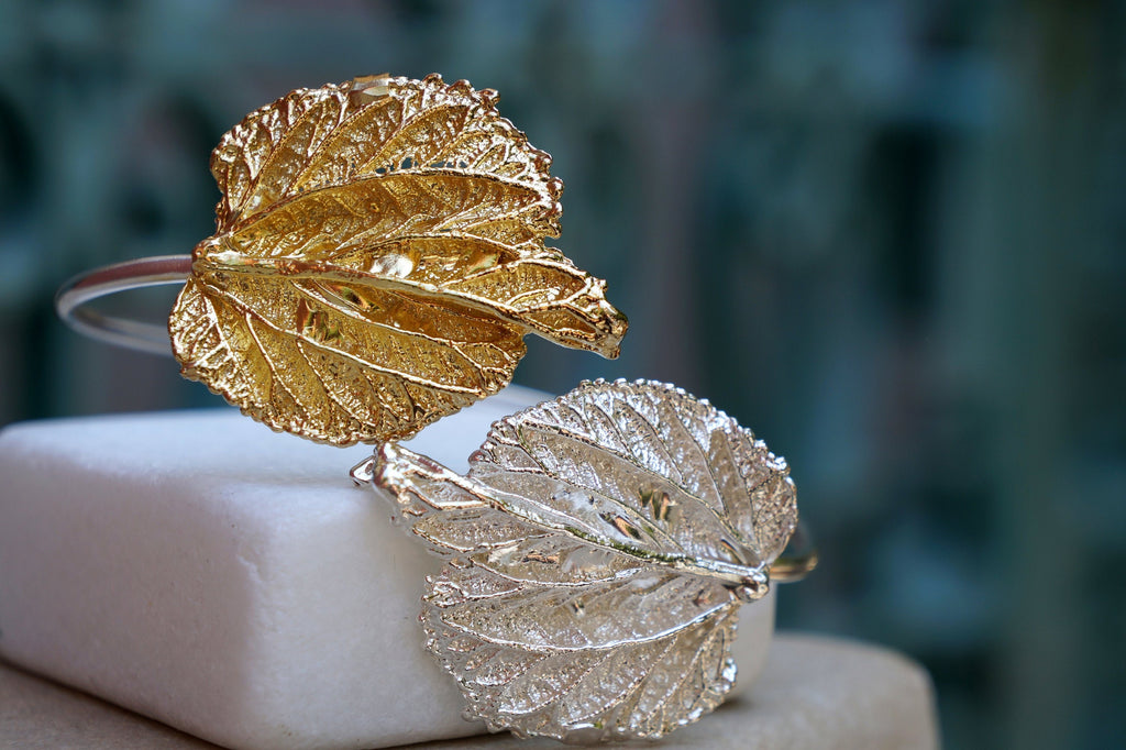Two ornate leaf-shaped bracelets, one gold and one silver, are displayed on a white surface against a blurred green background.