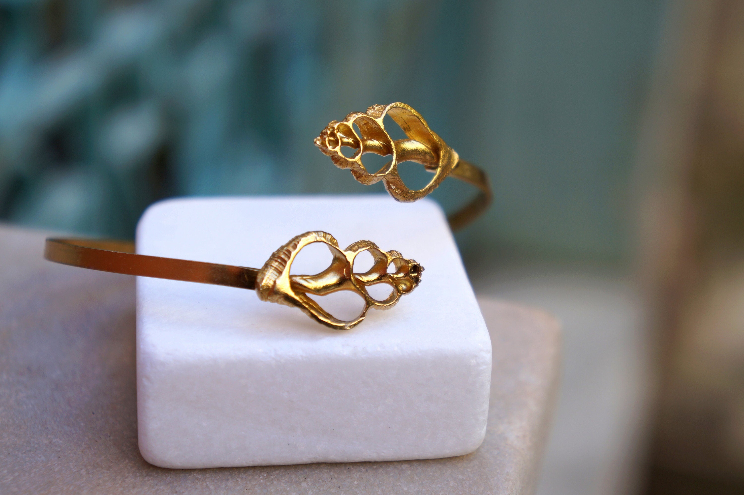 two gold-colored bracelets with intricate shell designs, placed on a white foam base.