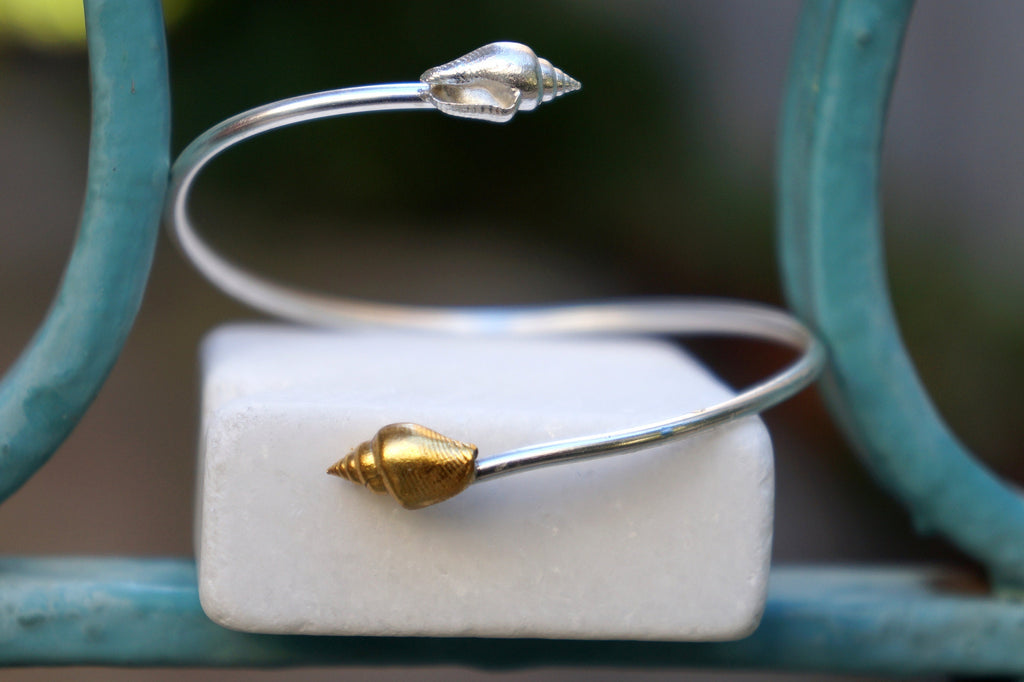 A silver and gold bracelet with a shell-shaped clasp is displayed on a white marble block, with a blurred green background.