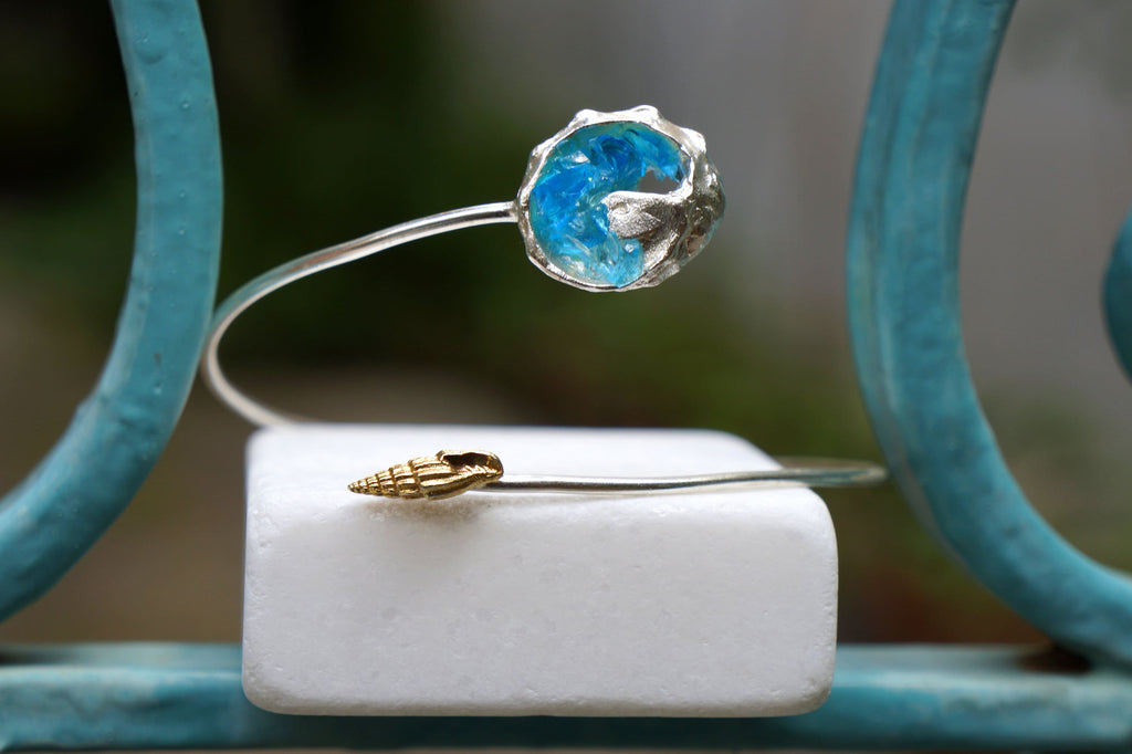 A silver bracelet with a blue gemstone and a gold seashell charm is displayed on a white marble surface.