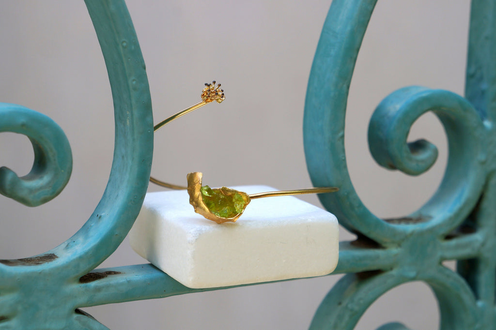 a piece of jewelry, specifically a bracelet, placed on a white square object. The bracelet is made of gold and has a green gemstone in the center. The bracelet is positioned on a blue metal railing, which adds a contrasting color to the scene.