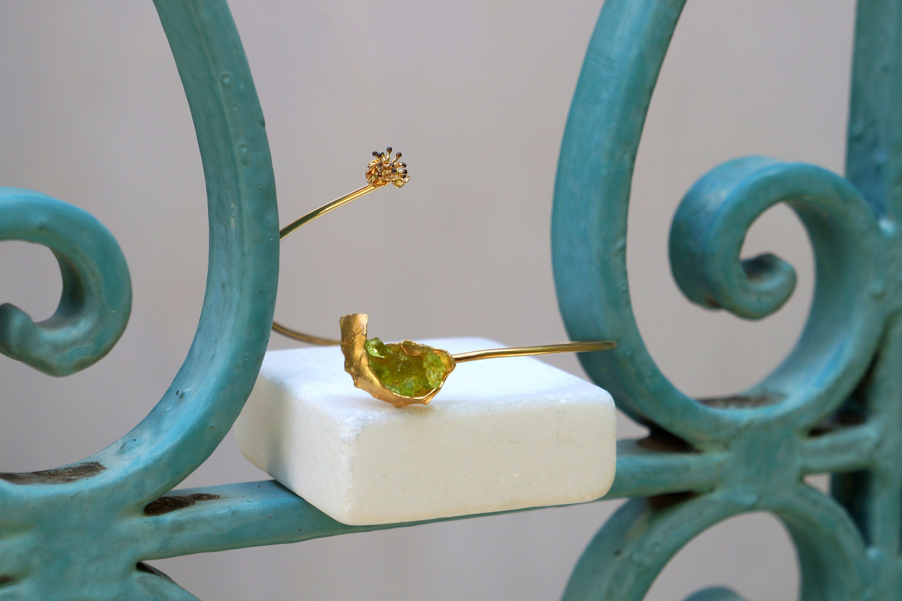 a piece of jewelry, specifically a bracelet, placed on a white square object. The bracelet is made of gold and has a green gemstone in the center. The bracelet is positioned on a blue metal railing, which adds a contrasting color to the scene.