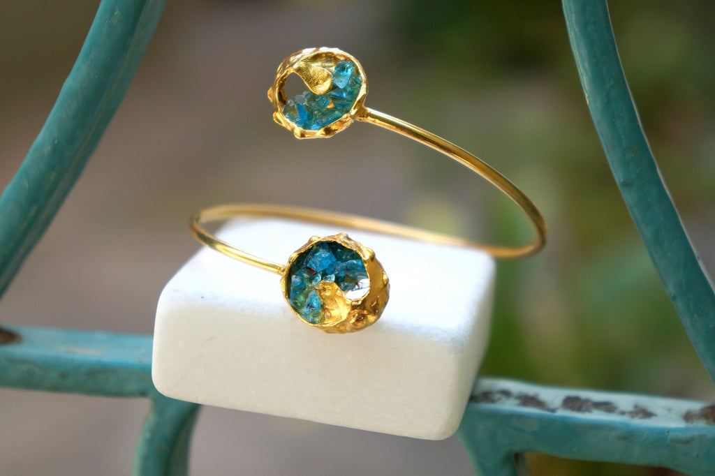 a gold bracelet with two round, blue gemstone-like stones attached to it, placed on a white square surface.