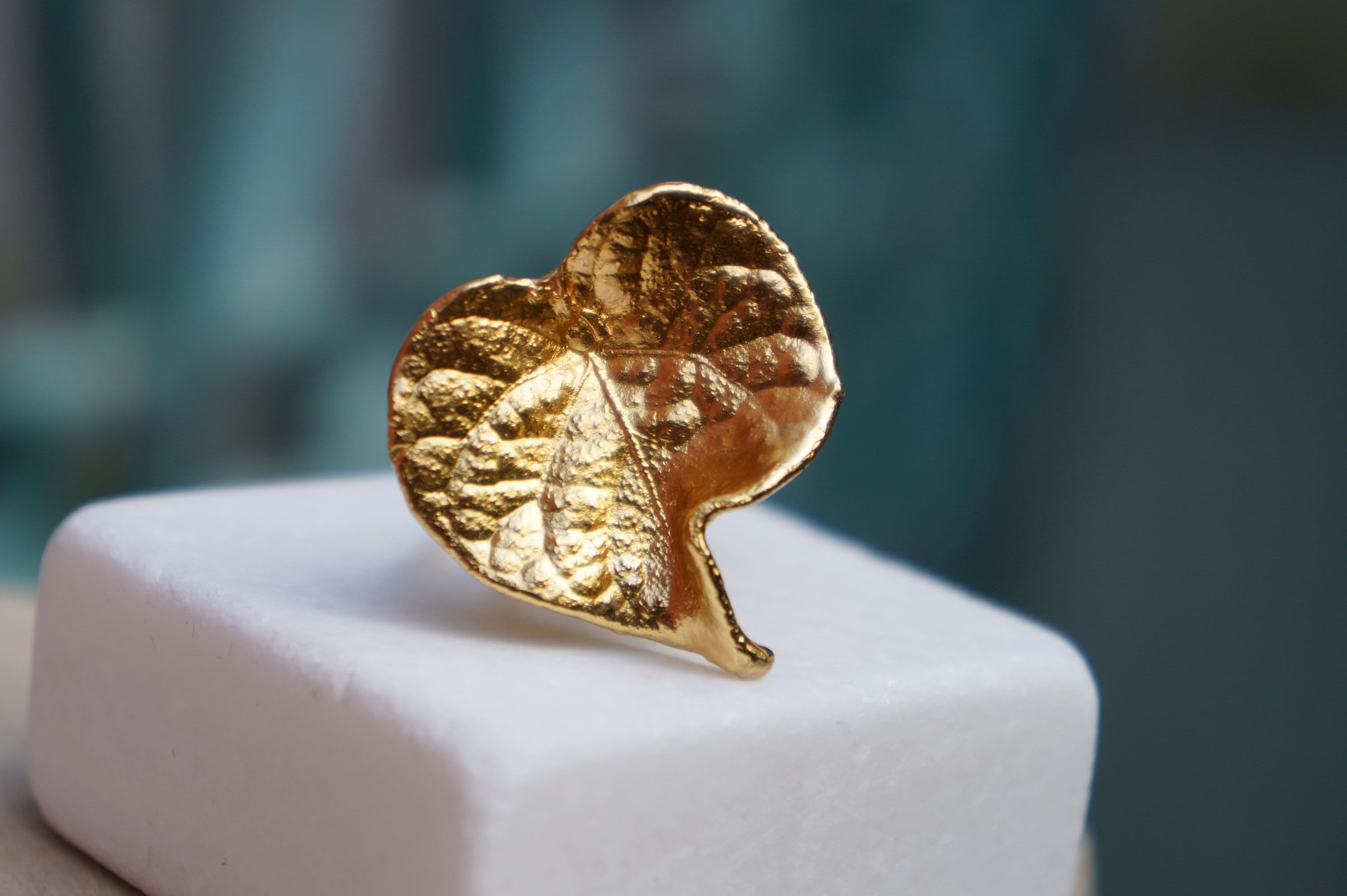 A close-up image of a gold leaf ring or pendant resting on a white surface.