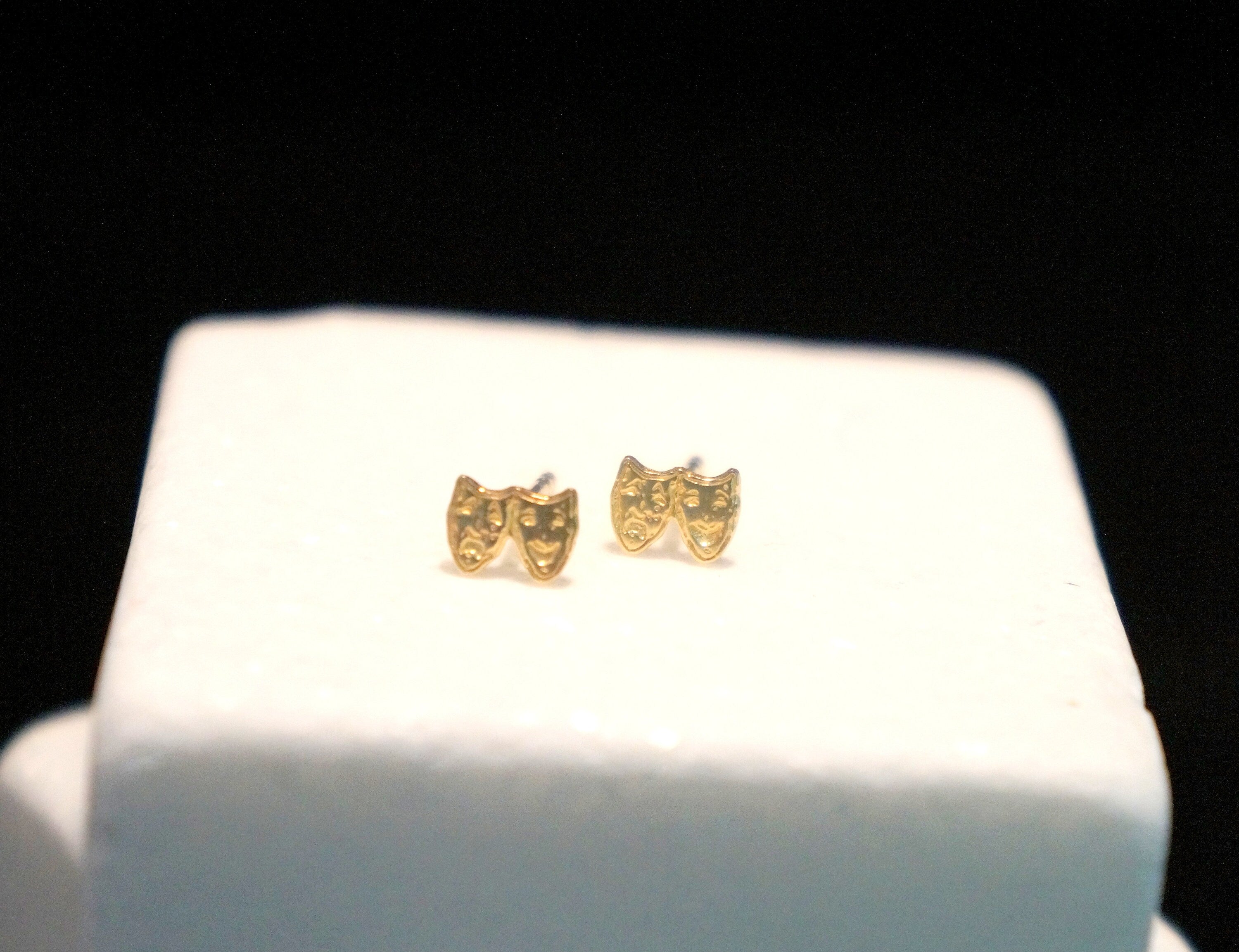 A pair of gold-colored earrings with theatrical masks on them, displayed on a white surface against a black background.