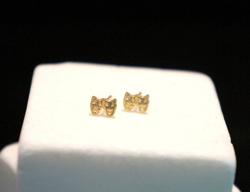 A pair of gold-colored earrings with theatrical masks on them, displayed on a white surface against a black background.