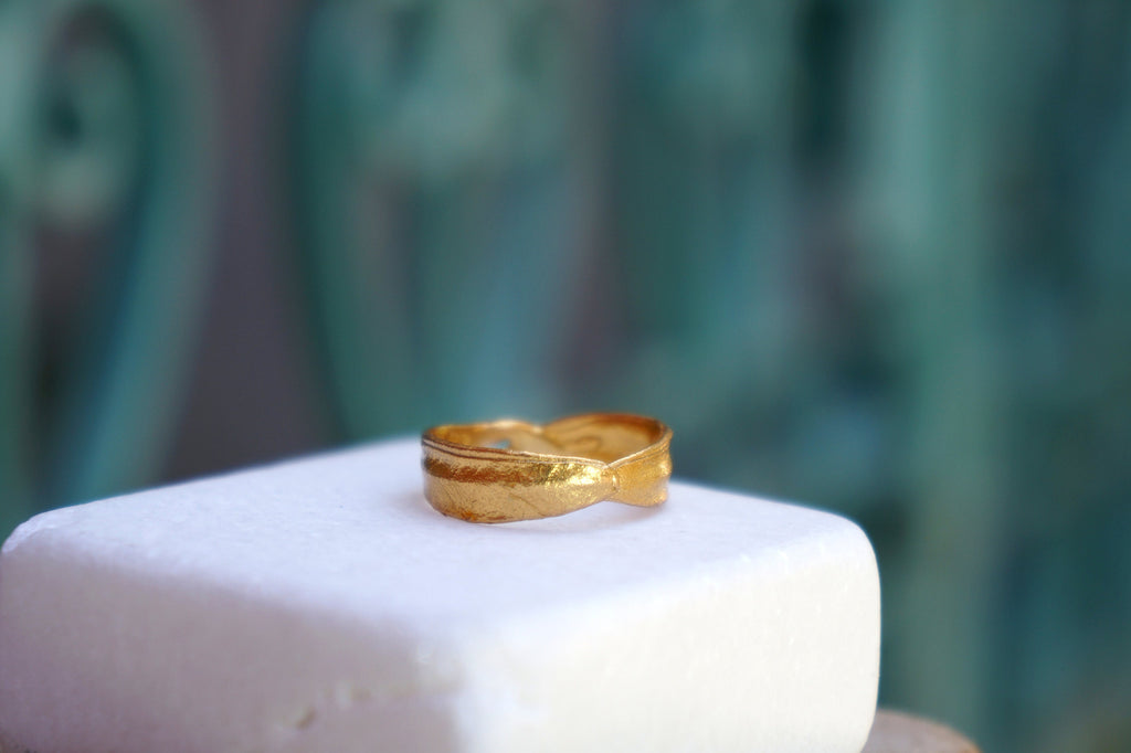 A gold ring with a bow-shaped band sits on a white square surface against a blurred green background.