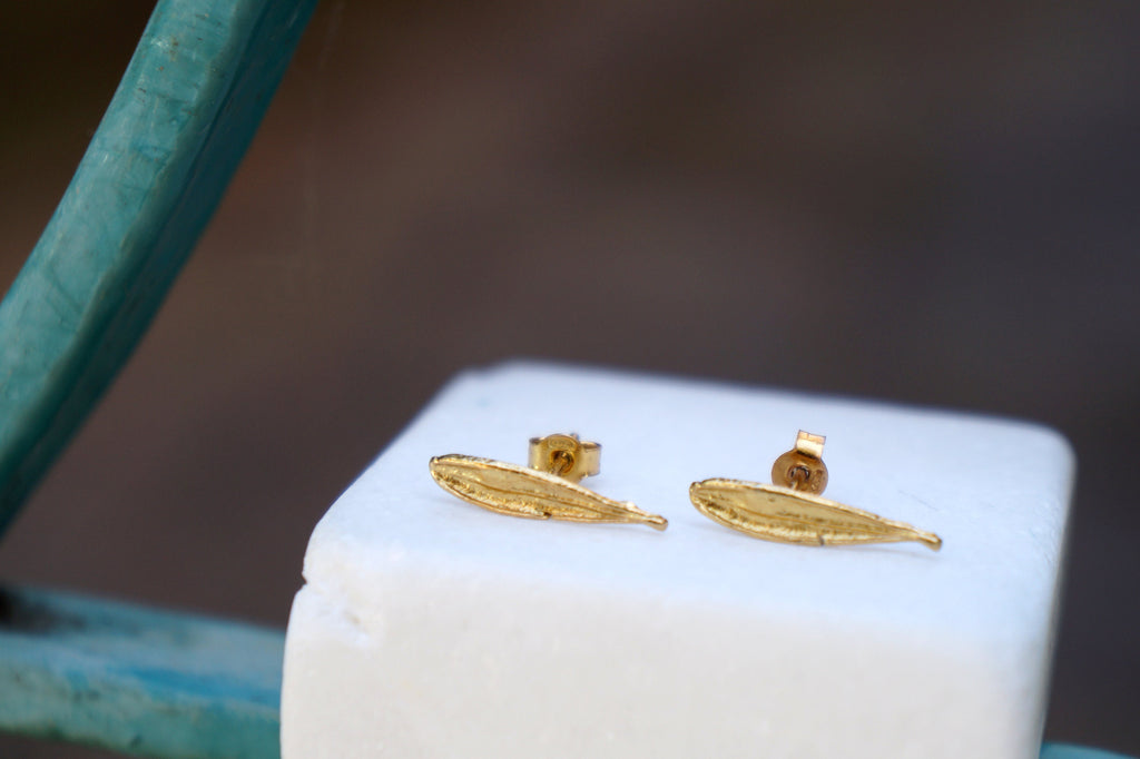 A pair of gold leaf earrings is displayed on a white pedestal against a blurred background.