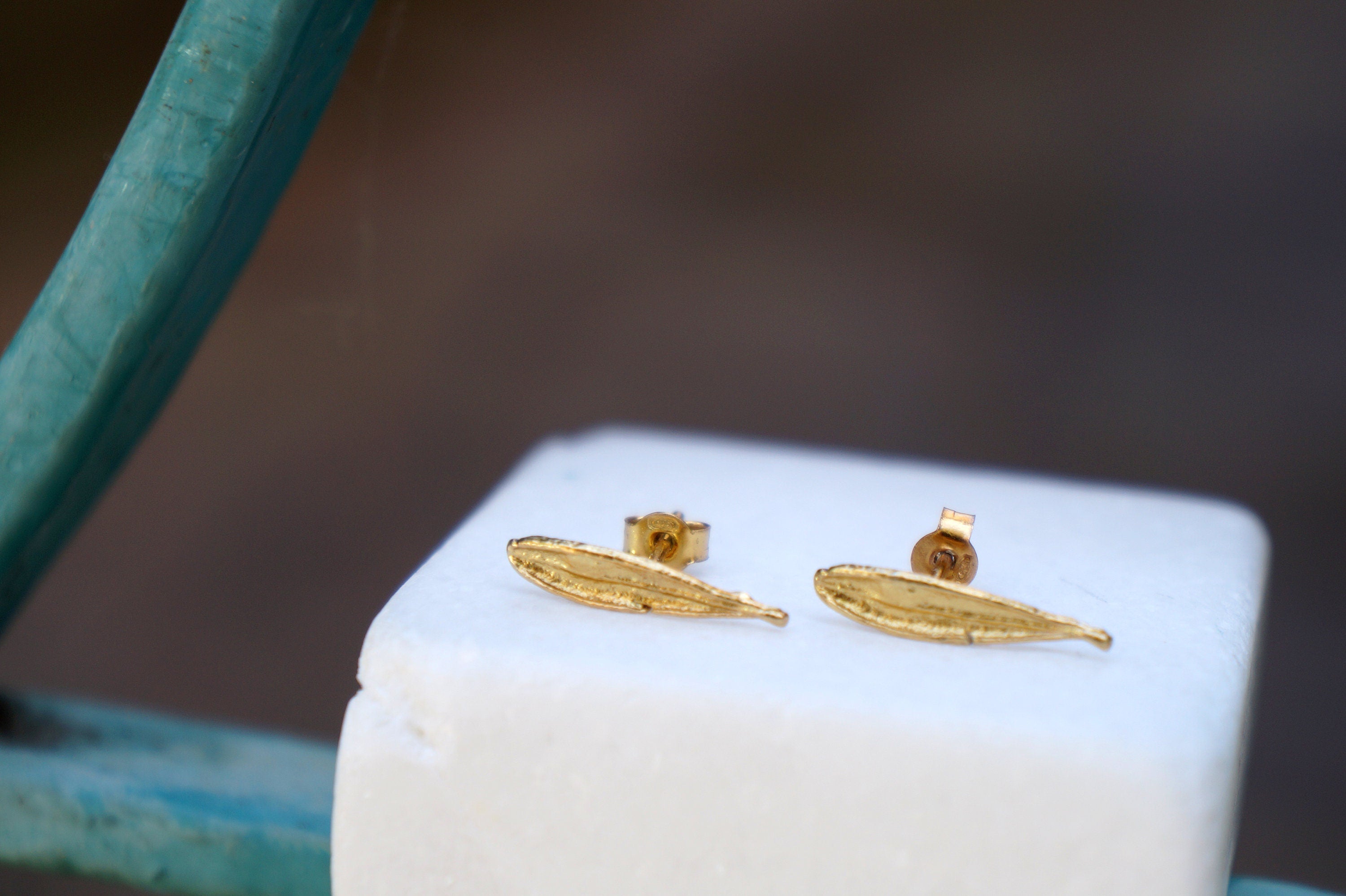 A pair of gold leaf earrings is displayed on a white pedestal against a blurred background.