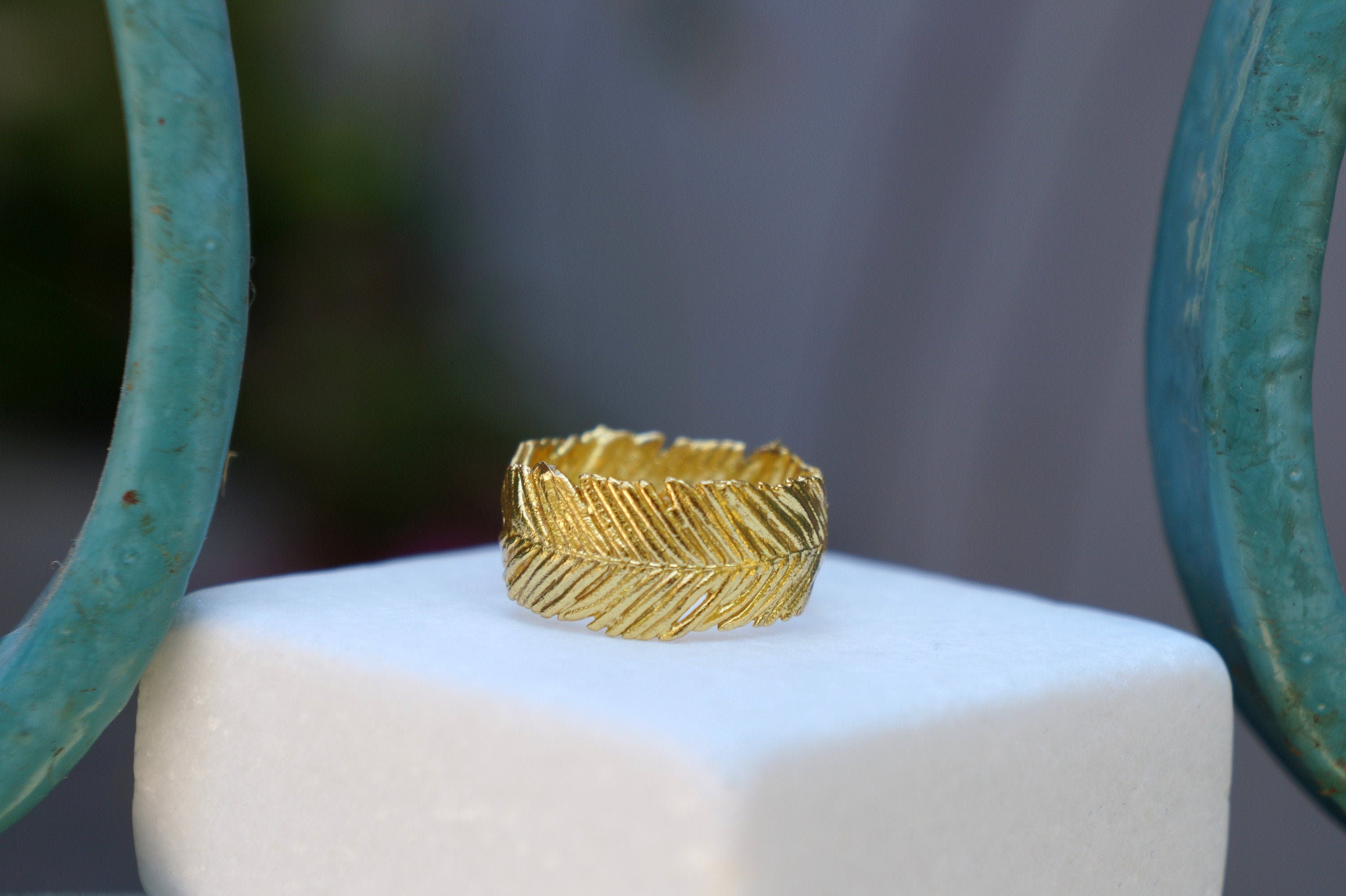 A gold ring with a leaf-like design sits on a white surface, with a teal-colored metal railing visible in the background.