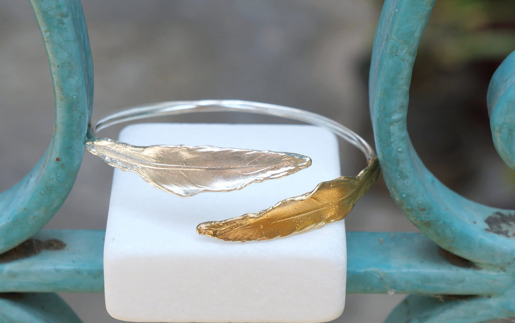 A silver bracelet with gold leaf-shaped charms is displayed on a white surface, with a blue metal railing in the background.