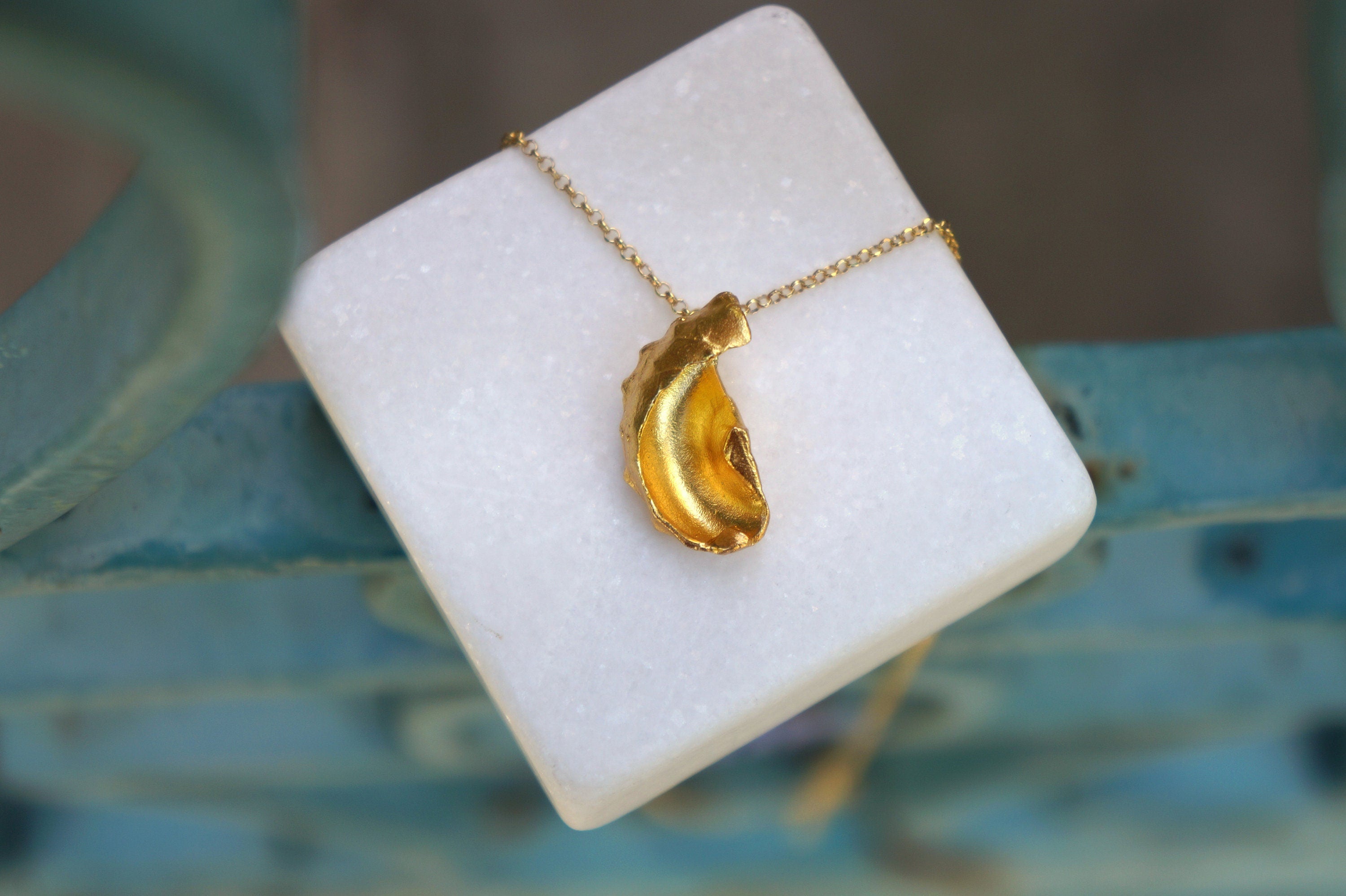 A gold-colored pendant necklace is displayed on a white square surface, with a blurred blue background.