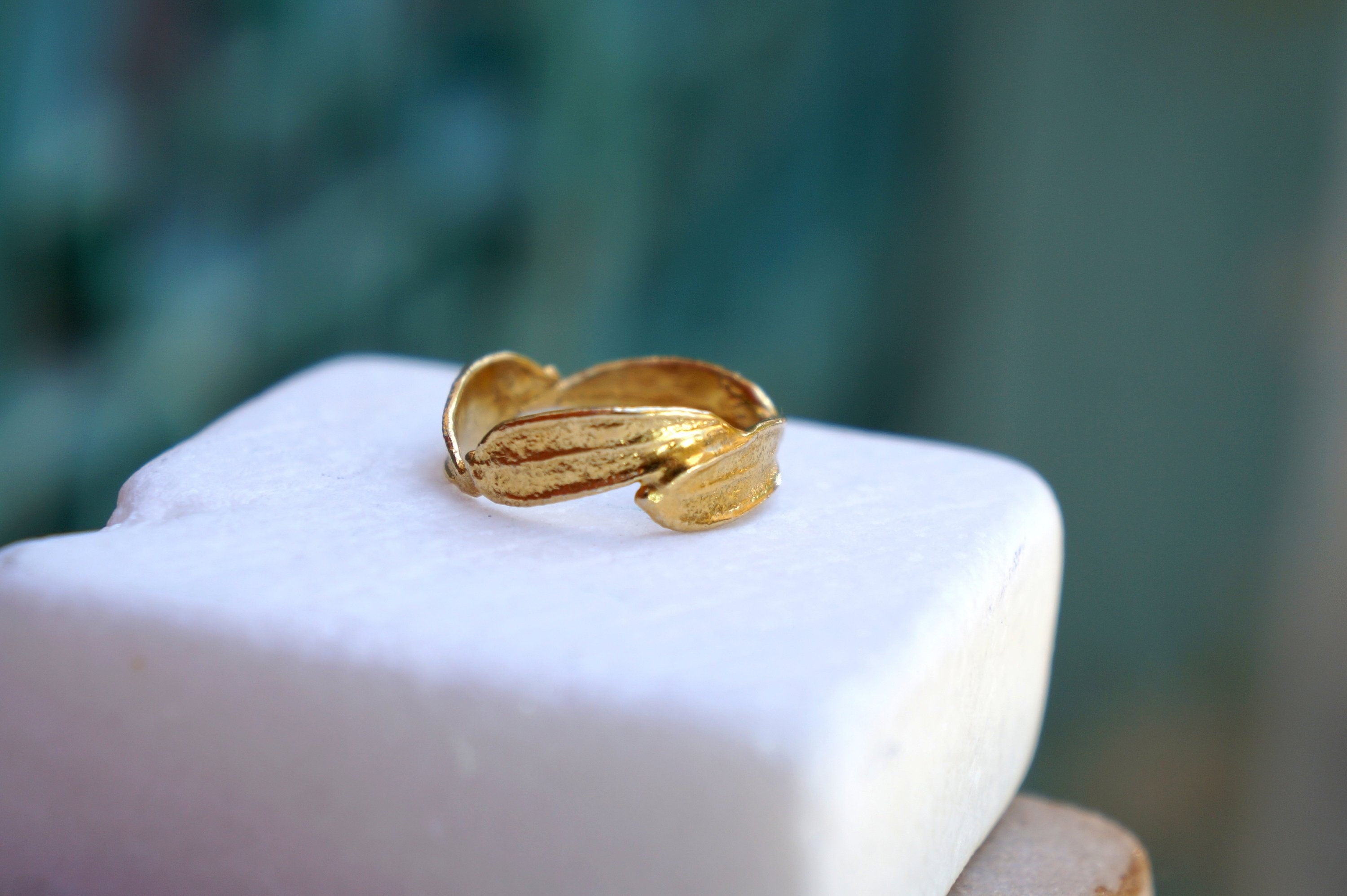 A gold ring with leaf-shaped designs sits on a white surface against a blurred green background.
