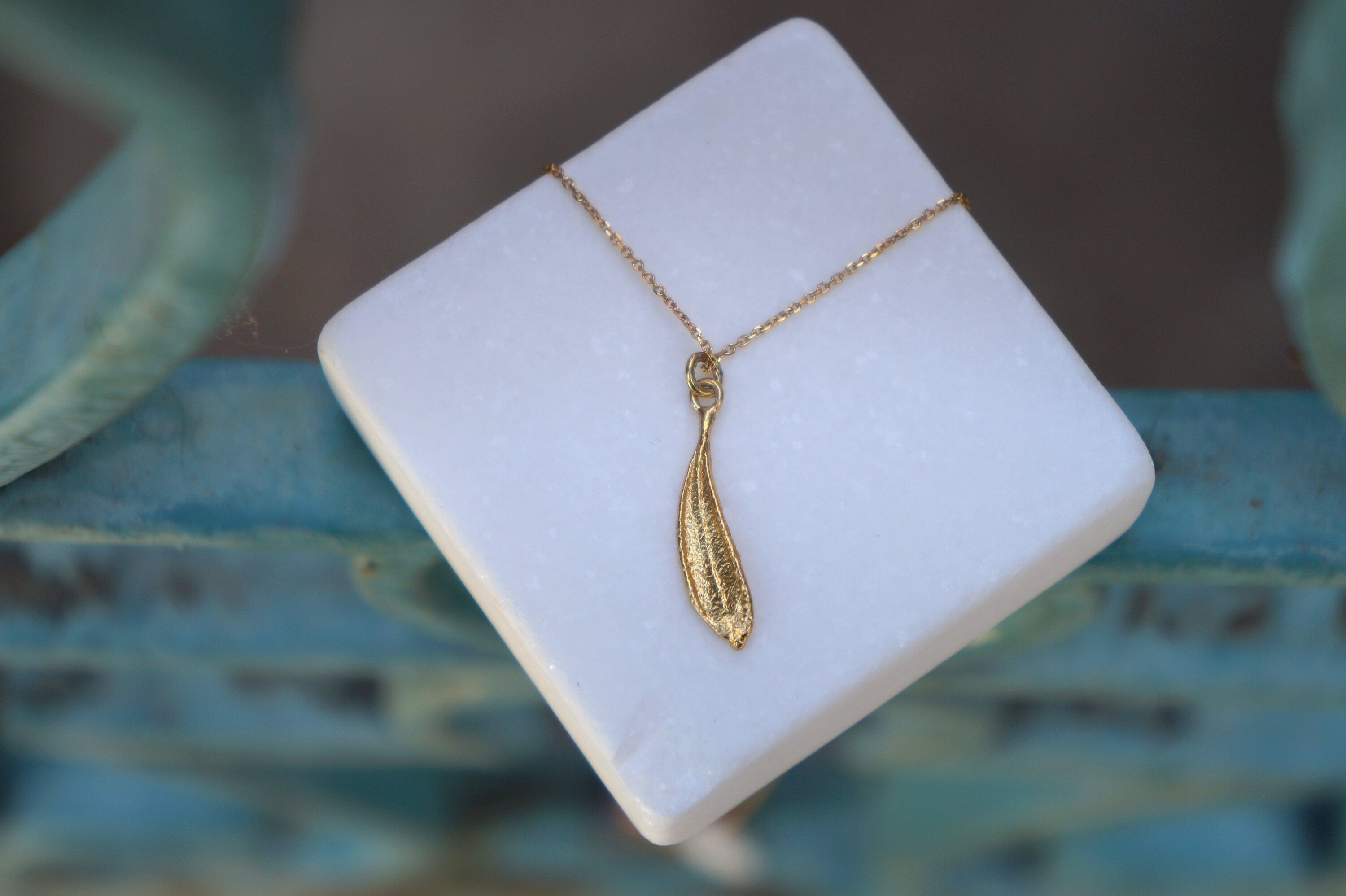 A gold-colored leaf pendant necklace is displayed on a white square surface, with a blurred blue background.