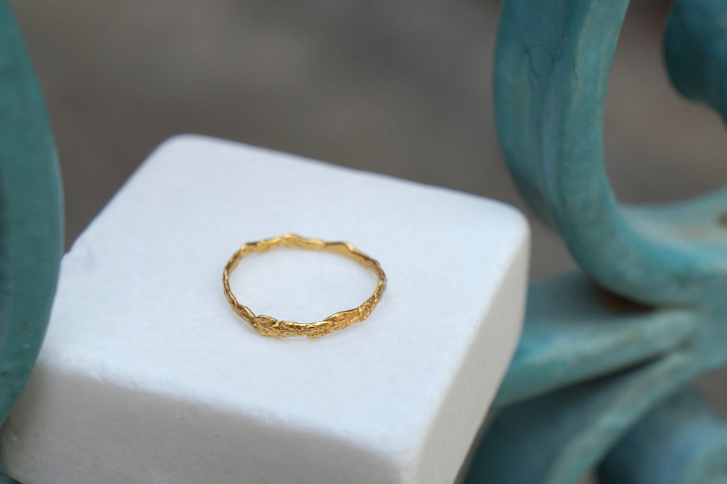 A gold ring with a leaf-like design rests on a white square surface, with a blurred teal-colored background.