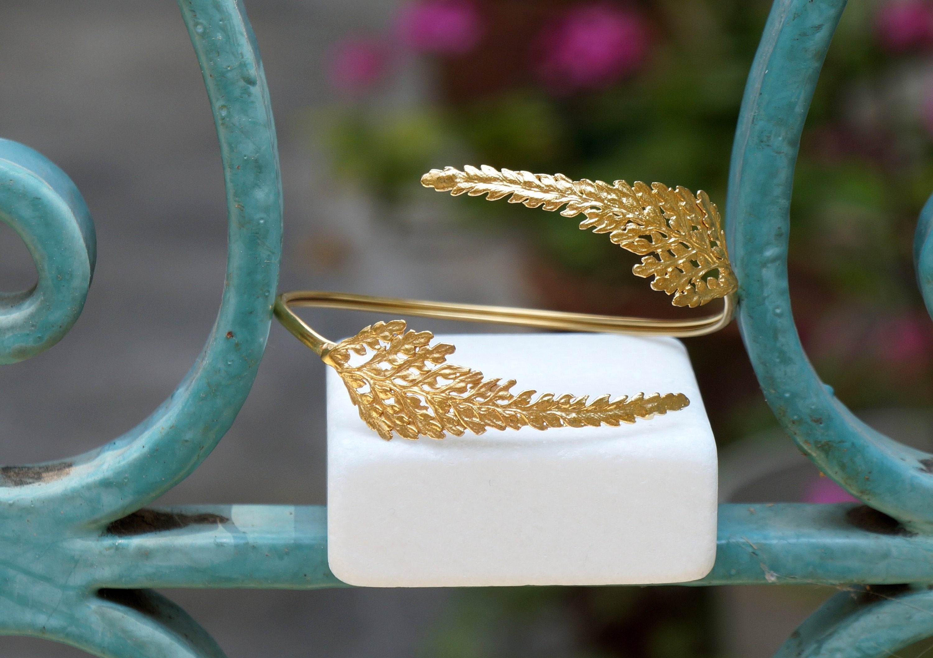 A gold bracelet with leaf-shaped designs is displayed on a white pedestal against a blurred background of flowers.