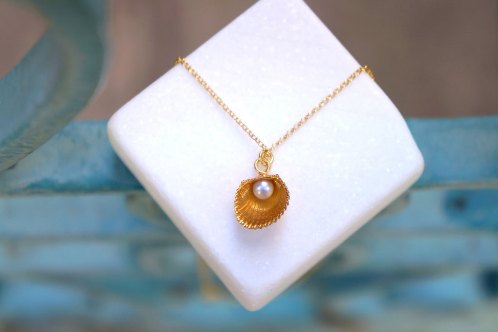 A gold necklace with a pearl pendant is displayed on a white square surface against a blue background.