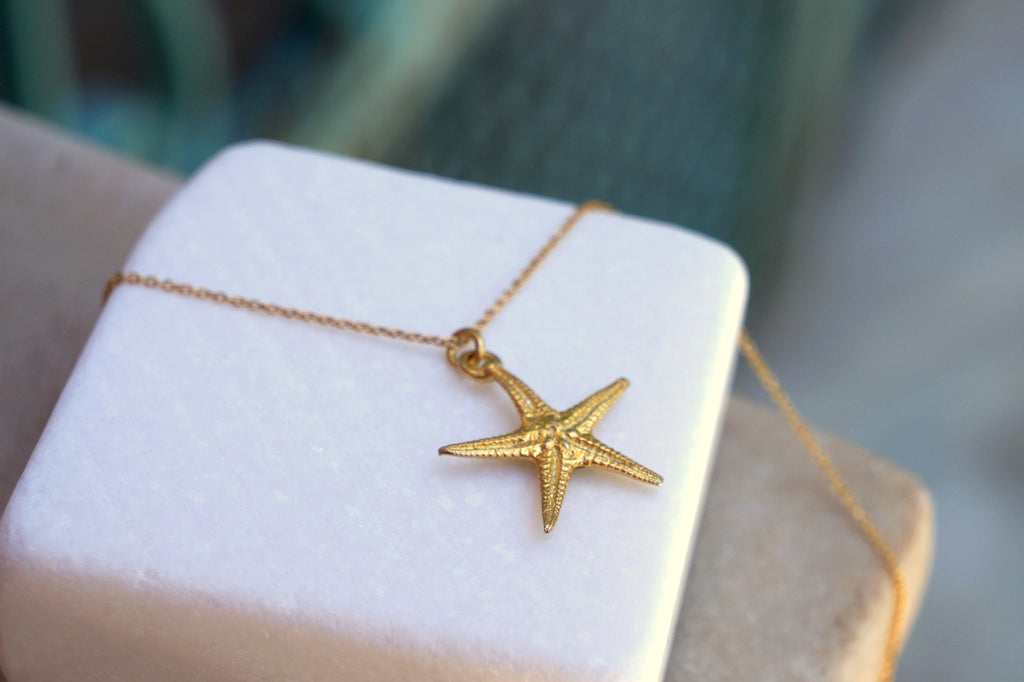 A gold starfish pendant is displayed on a white square box, with a gold chain necklace visible.