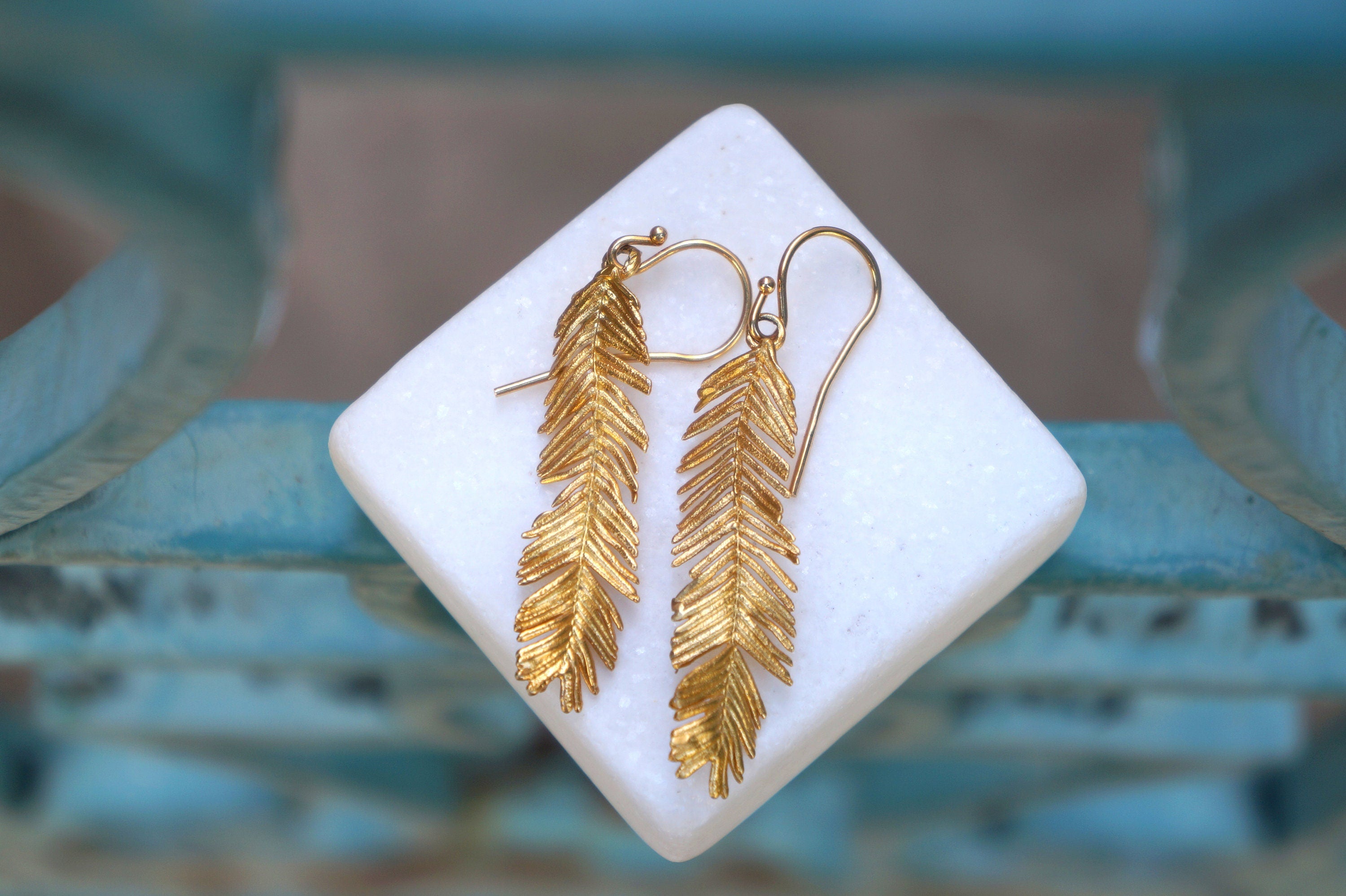 A pair of gold earrings with feather-like designs, displayed on a white square surface against a blurred blue background.