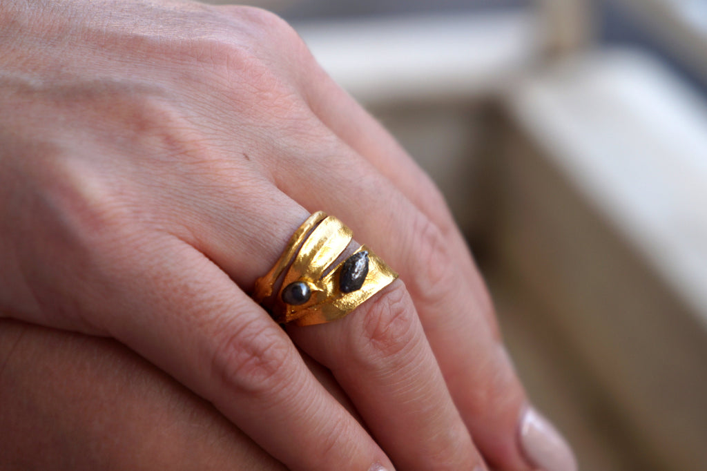 A close-up view of a person's hand wearing a gold ring with a leaf-shaped design and two black stones.