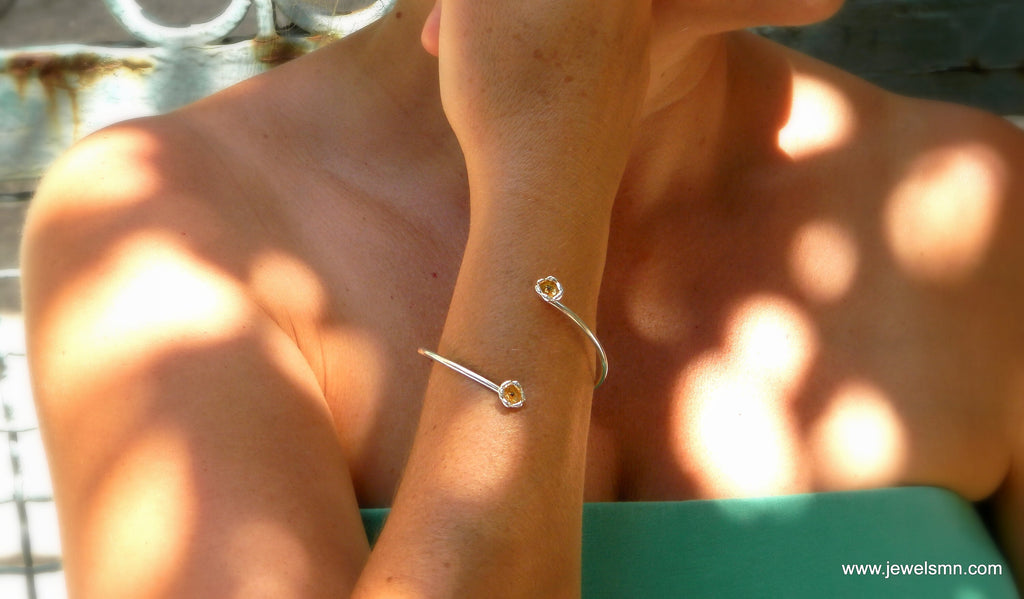 A close-up of a person's arm wearing a silver bracelet with two small charms, with sunlight casting a pattern on their skin.