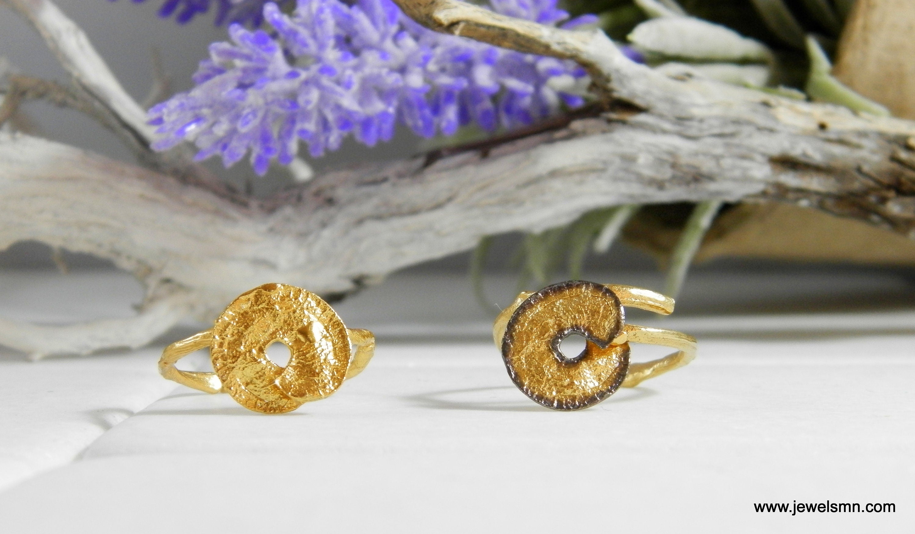 Two gold rings with unique designs are placed on a white surface, with a branch of purple flowers and a wooden branch in the background.