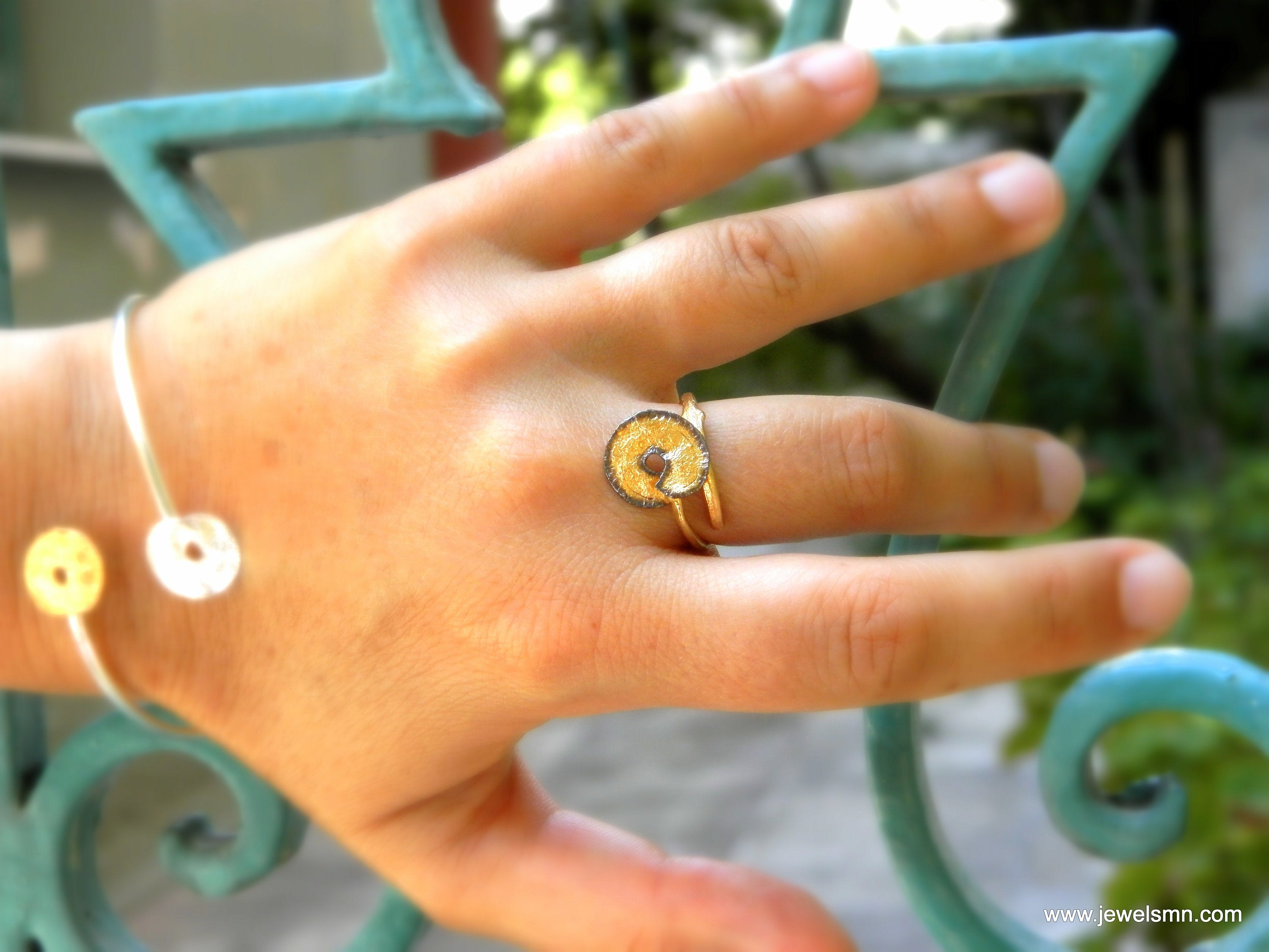 A close-up of a person's hand wearing a gold ring and bracelet, with a blurred background of greenery.