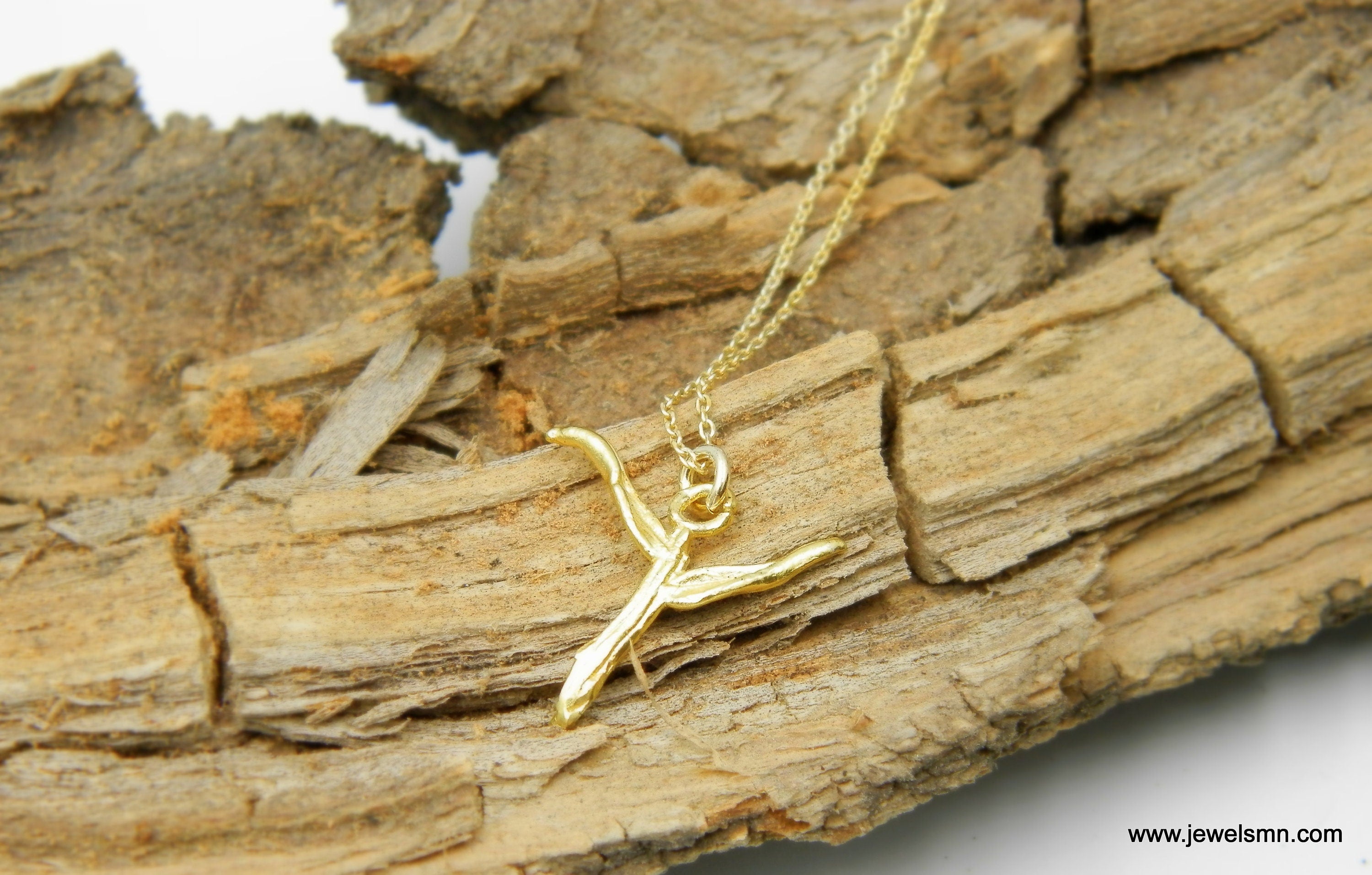 A gold-colored pendant in the shape of a letter 'X' is resting on a piece of weathered wood.