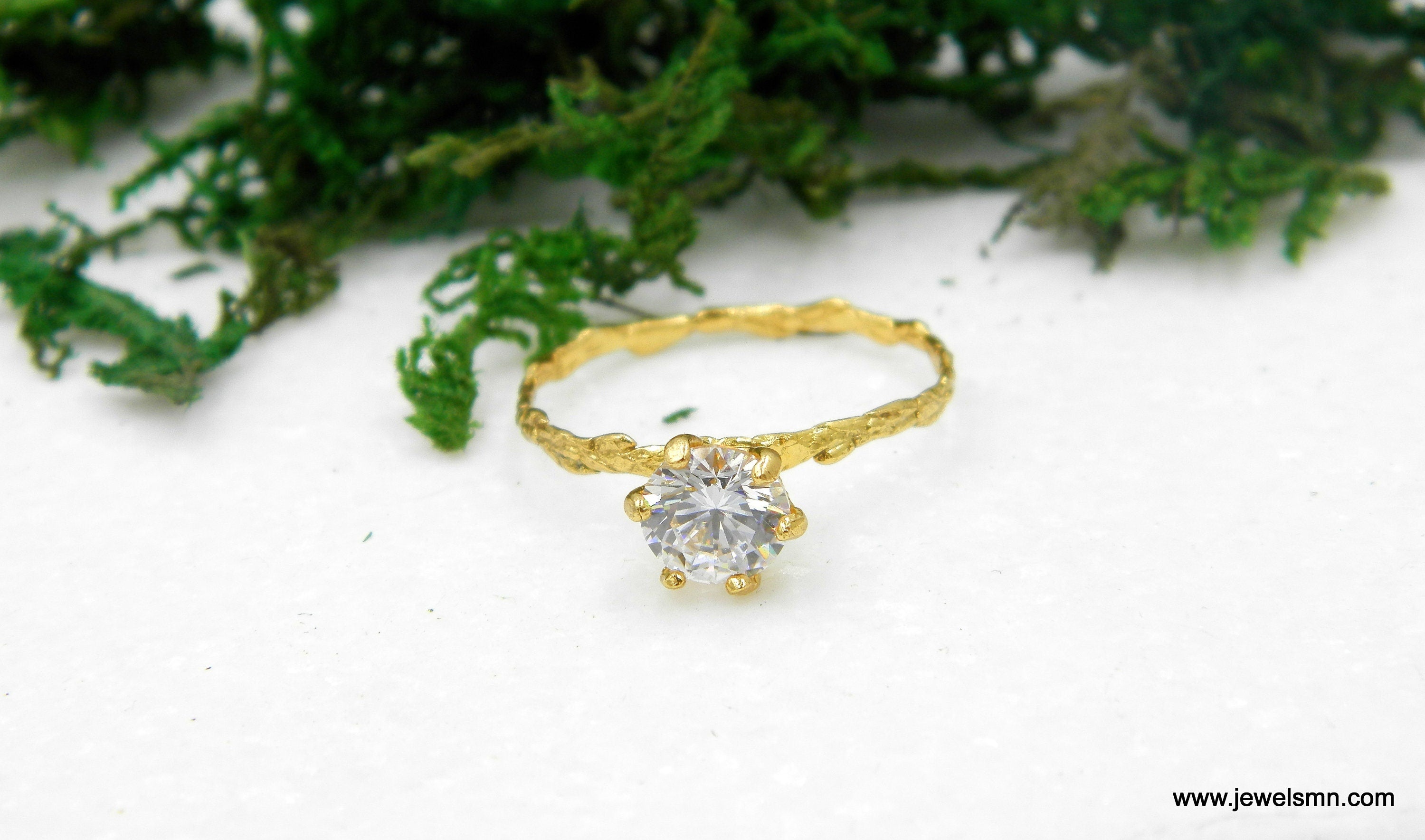 A gold ring with a large diamond set in the center, placed on a white surface with green foliage in the background.