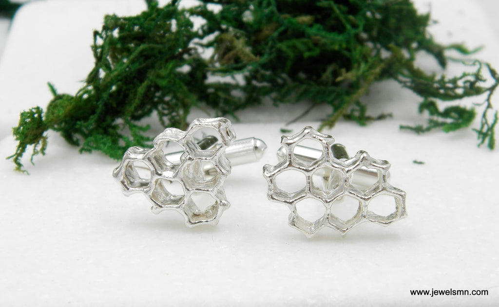 two silver cufflinks with a honeycomb design, placed on a white surface with a green mossy background.