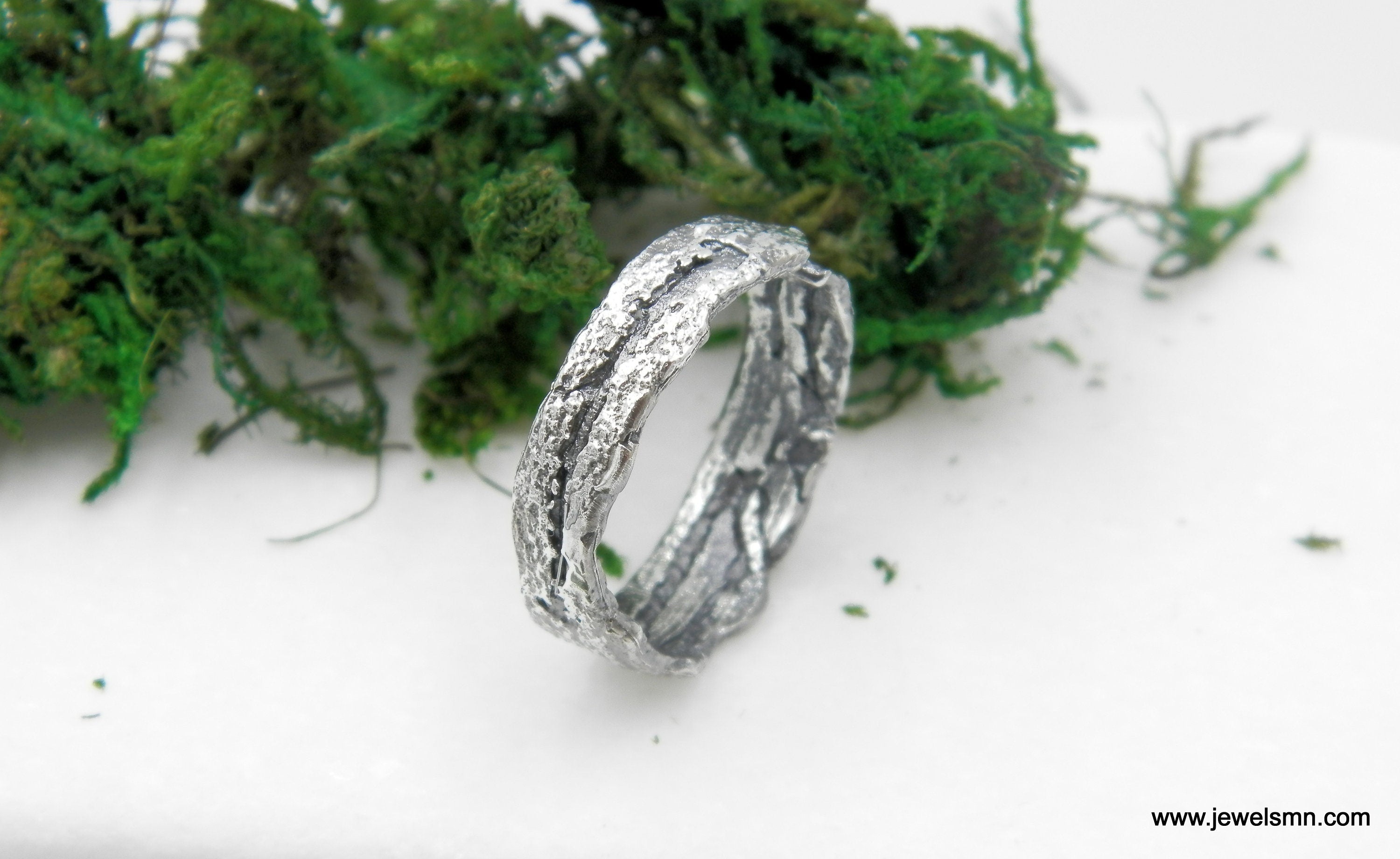 A silver ring with a unique, textured design sits on a white surface, surrounded by green moss.