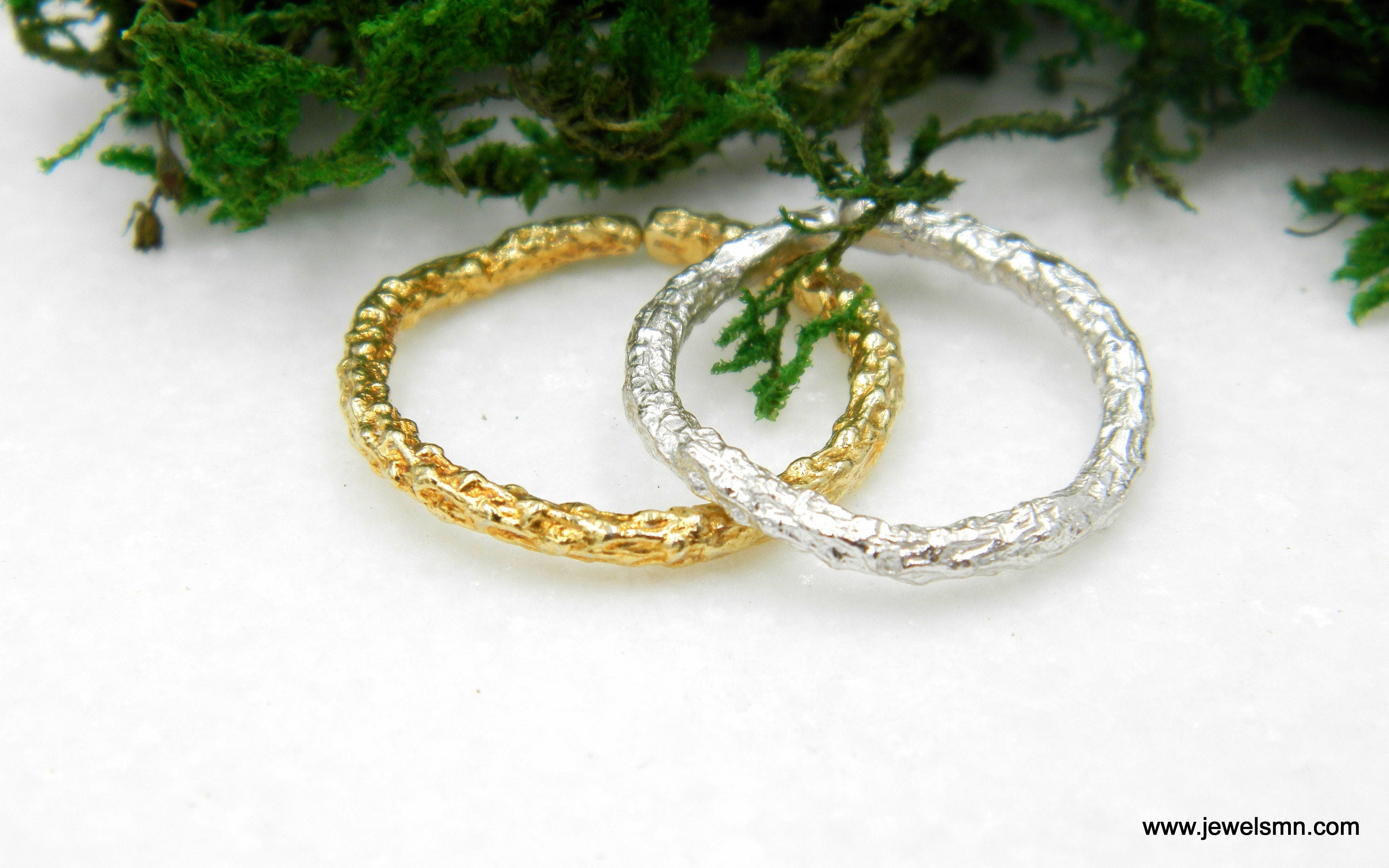 Two rings, one gold and one silver, are placed on a white surface with green foliage in the background.