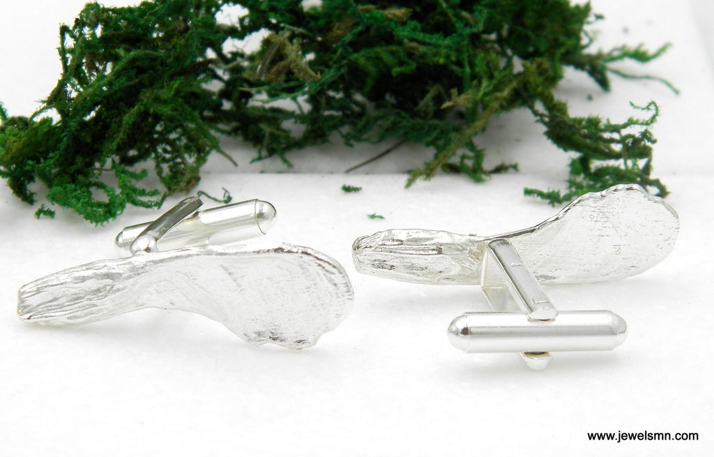 a pair of silver cufflinks with a leaf-like design, placed on a white surface next to a pile of green moss.