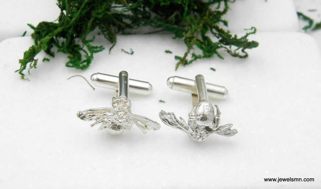 two silver cufflinks with bee-like designs, placed on a white surface with some green moss.