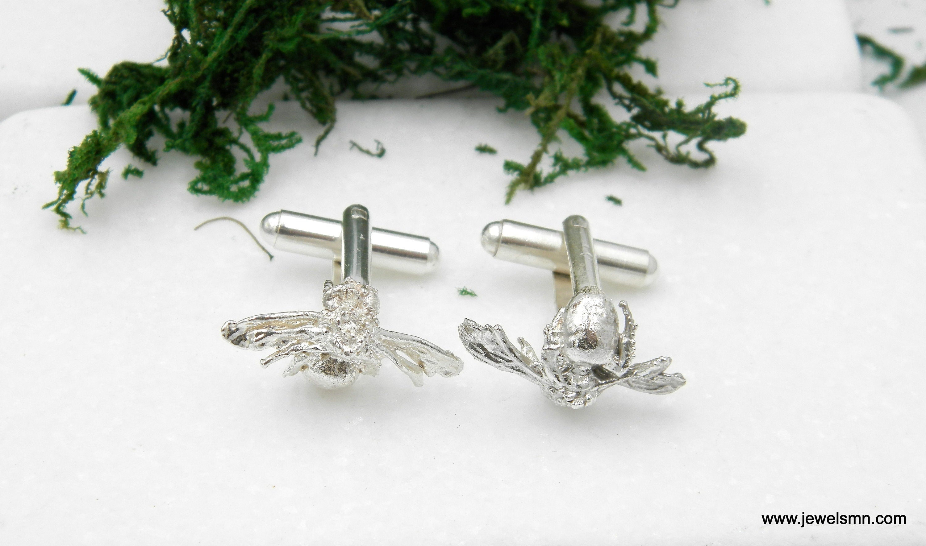 two silver cufflinks with bee-like designs, placed on a white surface with some green moss.
