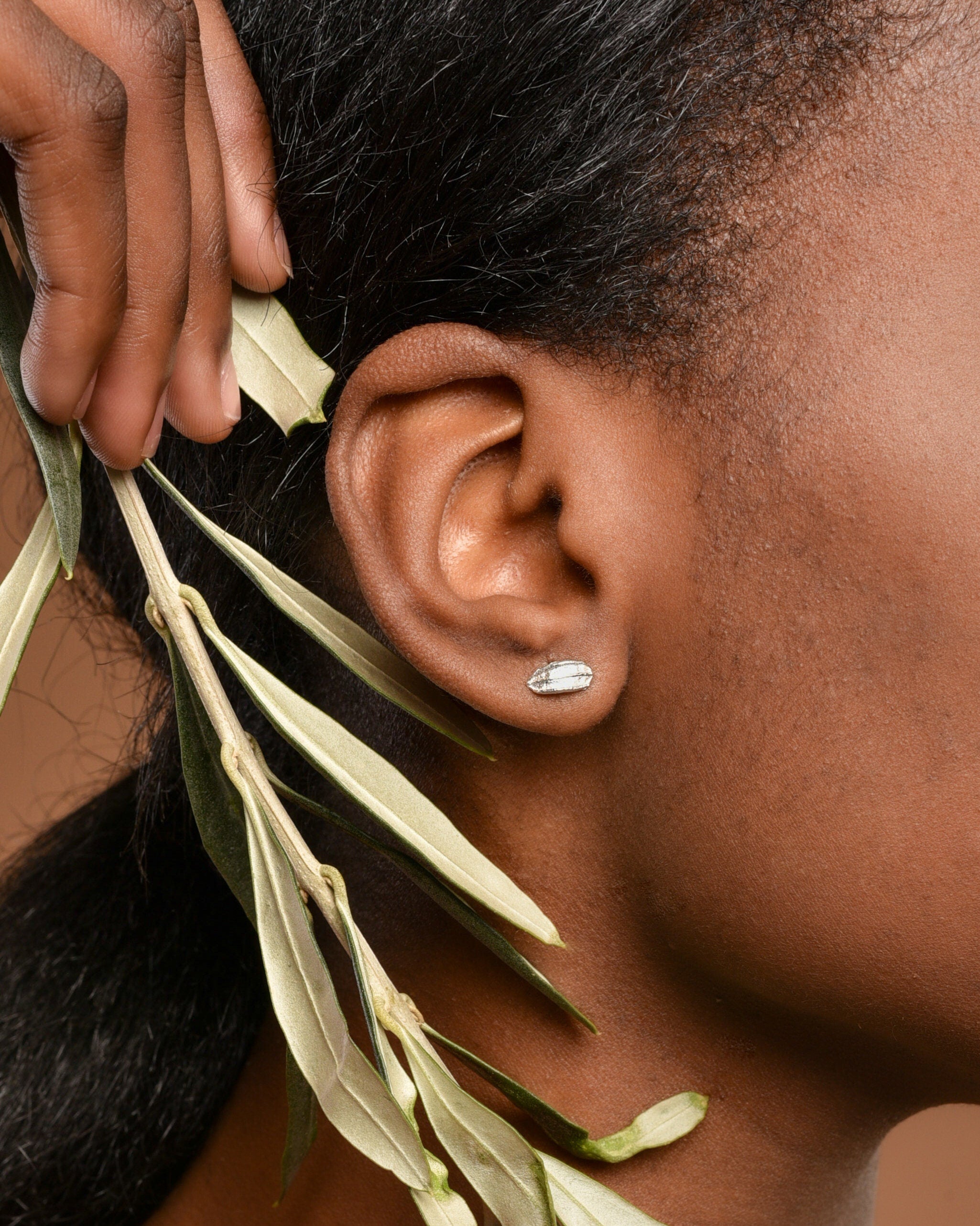 a close-up view of a person's ear adorned with a small earring, with a branch of green leaves partially obscuring the ear.
