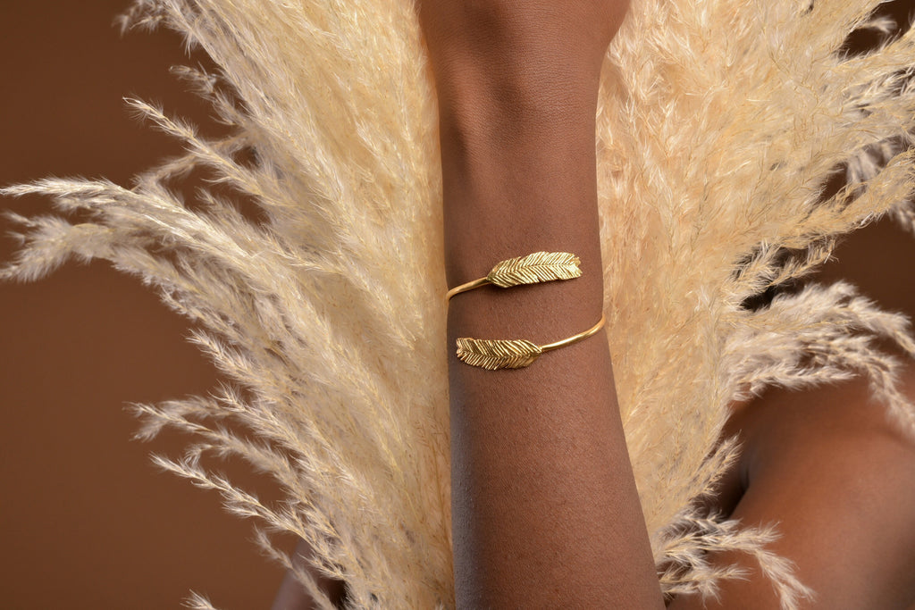 A gold bracelet with two feather-shaped charms is worn on a person's wrist.
