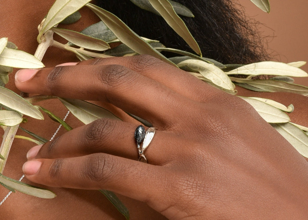 A close-up of a hand wearing a silver ring with a leaf-like design, resting on a branch of green leaves.
