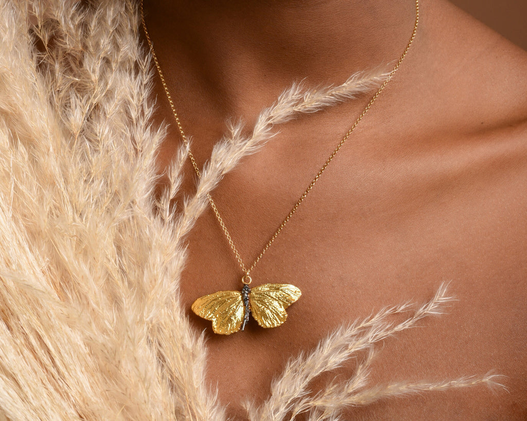 A close-up of a gold butterfly pendant necklace worn around a person's neck, with a beige feather-like fabric draped over the neck.