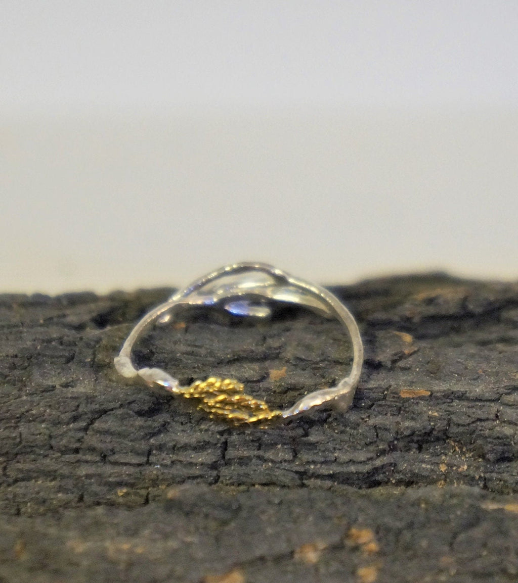 A silver ring with a gold leaf design rests on a dark, textured surface.