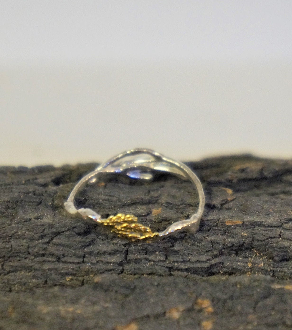 A silver ring with a gold leaf design rests on a dark, textured surface.