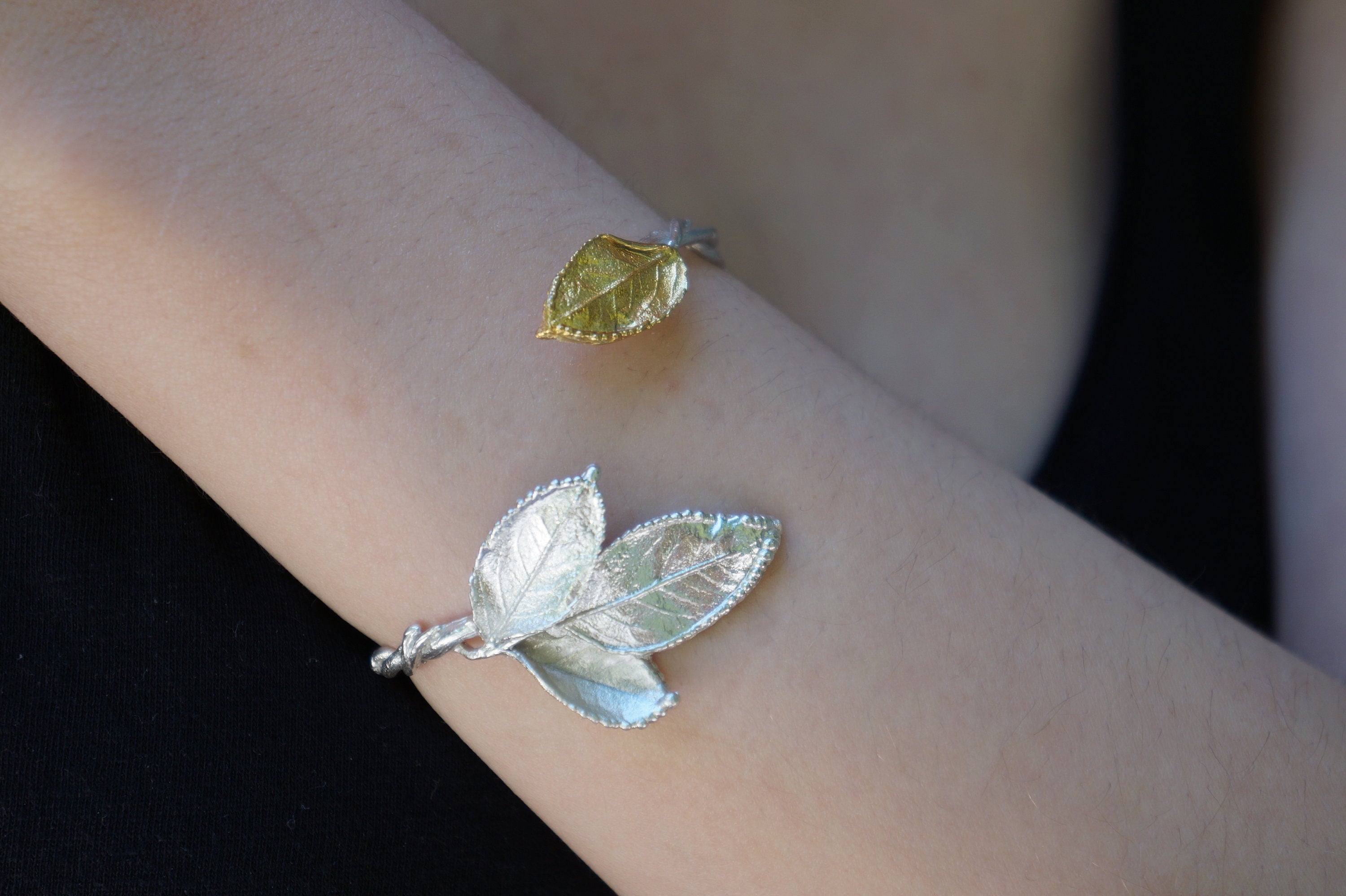 A person's arm wearing a bracelet with a leaf-shaped charm and a ring with a leaf-shaped charm.