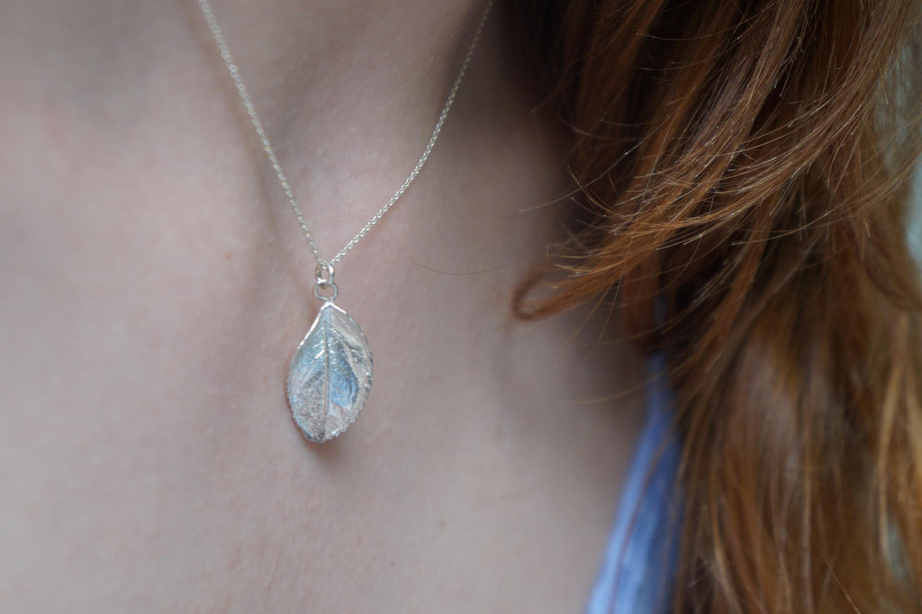A silver leaf pendant necklace is worn by a person with long, reddish-brown hair.