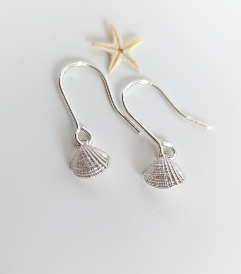 A pair of silver earrings with seashell-shaped charms, placed on a white surface alongside a small starfish.