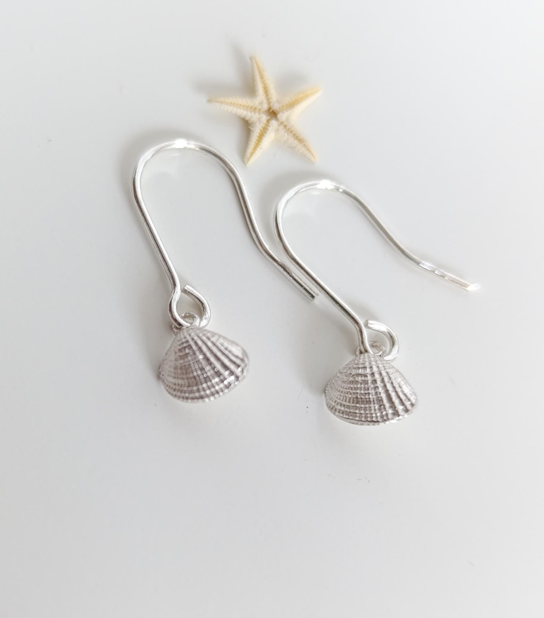 A pair of silver earrings with seashell-shaped charms, placed on a white surface alongside a small starfish.