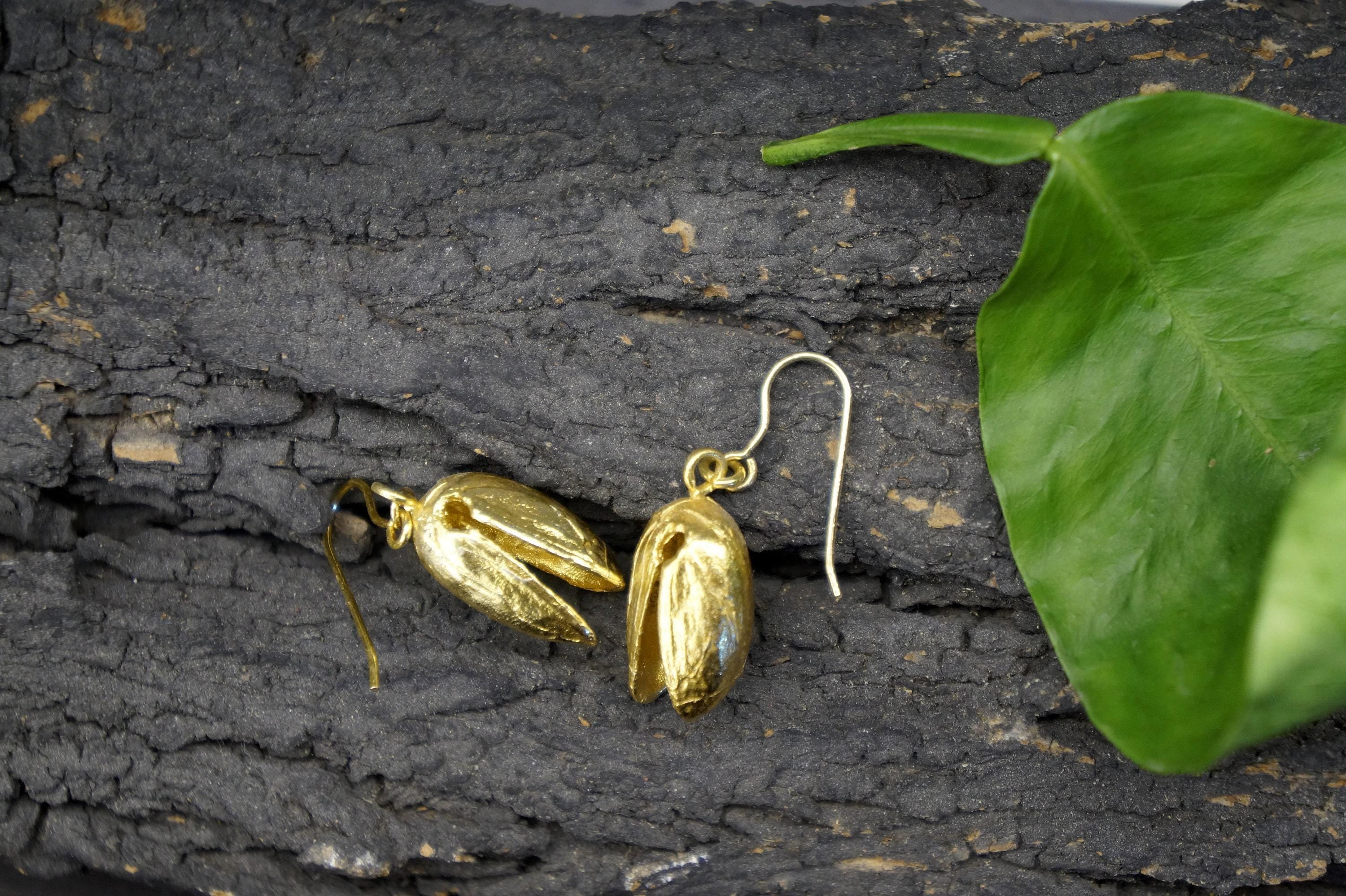 A pair of gold earrings with a leaf-like design rests on a textured surface, accompanied by a green leaf.