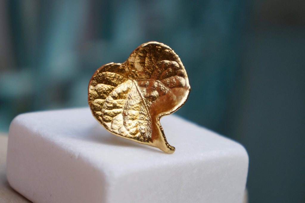 A close-up image of a gold leaf-shaped ring or brooch resting on a white surface.