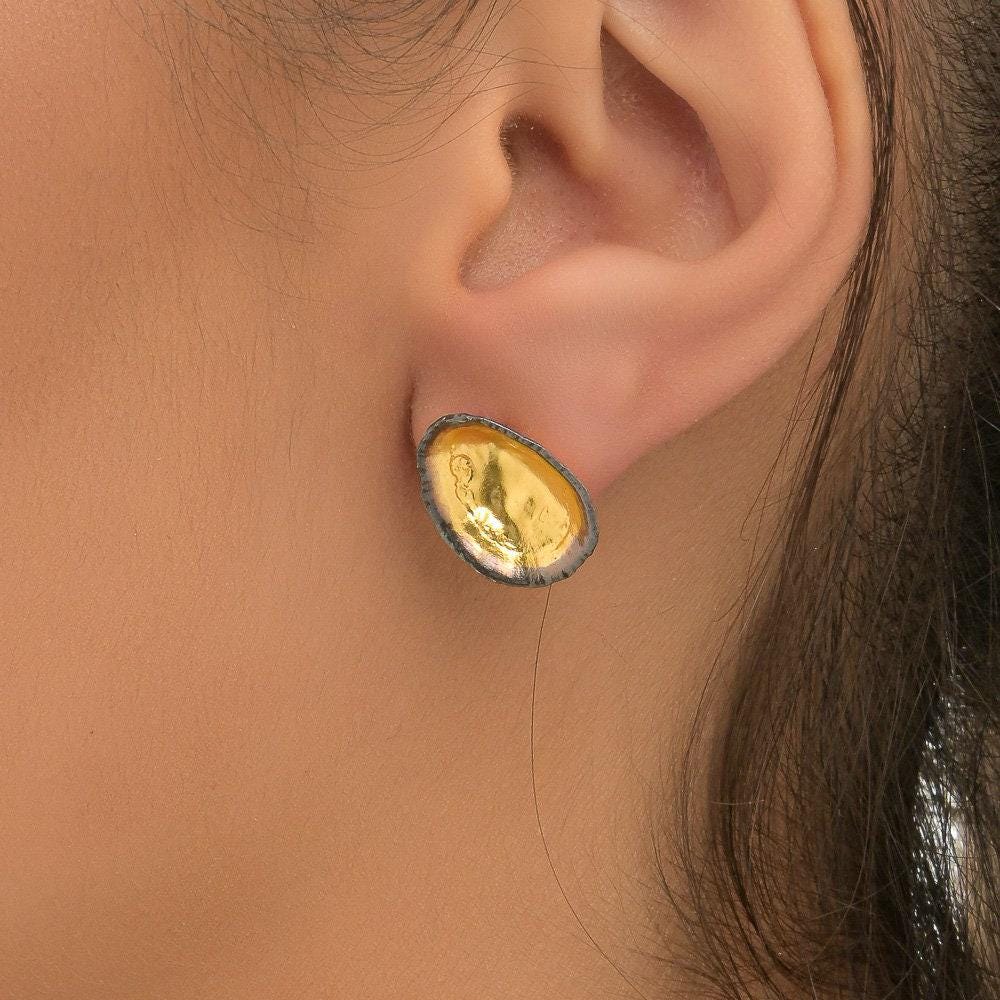 A close-up view of a person's ear wearing a pair of gold earrings with a unique, oval-shaped design.