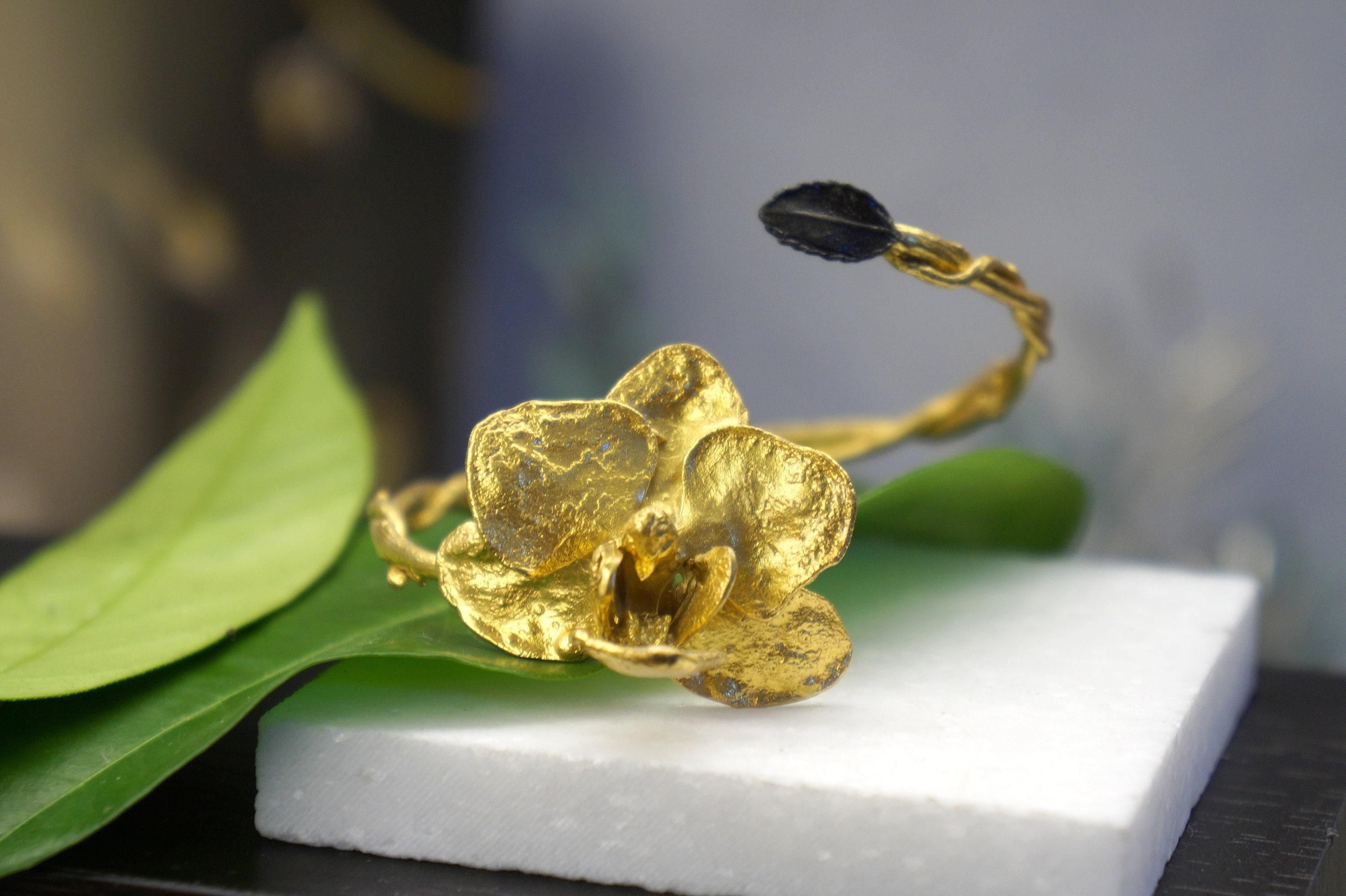 A close-up image of a golden orchid-shaped bracelet with a leaf-like design, resting on a white surface.